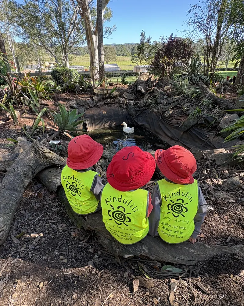 Three children sitting on a log watching a duck on a pond