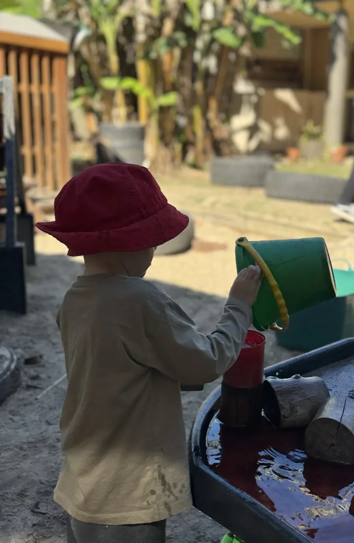 A child pouring water from a green bucket at the mud kitchen